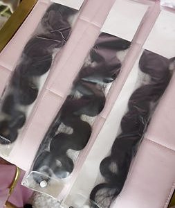 Long, black, wavy hair extensions in plastic packaging on a pink surface.